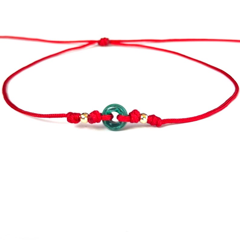 Puede incluir: Una pulsera de hilo rojo con una cuenta verde circular y dos peque&ntilde;as cuentas doradas. La pulsera est&aacute; anudada y es ajustable. El dise&ntilde;o es sencillo. El fondo es blanco.