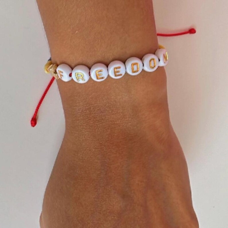 Peut inclure: Un bracelet en perles blanches avec le mot "FREEDOM" &eacute;crit en lettres dor&eacute;es. Le bracelet est attach&eacute; avec un cordon rouge et jaune.