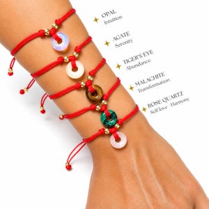 Puede incluir: Cinco pulseras de cordón rojo con dijes circulares de piedras preciosas que incluyen ópalo, ágata, ojo de tigre, malaquita y cuarzo rosa. Cada pulsera incluye pequeñas cuentas doradas y etiquetas de texto que identifican las piedras.