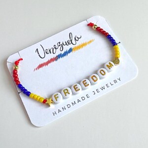 Peut inclure: Un bracelet en perles blanches avec des lettres dor&eacute;es qui &eacute;pellent "FREEDOM" et un charme d'&eacute;toile dor&eacute;e. Le bracelet est enfil&eacute; avec des perles rouges, bleues et jaunes, repr&eacute;sentant les couleurs du drapeau v&eacute;n&eacute;zu&eacute;lien. Le bracelet est pr&eacute;sent&eacute; sur une carte blanche avec le mot "Venezuela" &eacute;crit en cursive noire.