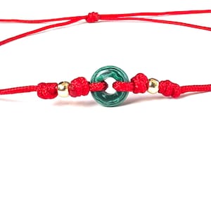 Puede incluir: Una pulsera de hilo rojo con una piedra verde circular con un patr&oacute;n &uacute;nico. La pulsera incluye cuentas doradas y nudos, sobre un fondo blanco. La cuerda est&aacute; anudada a intervalos.