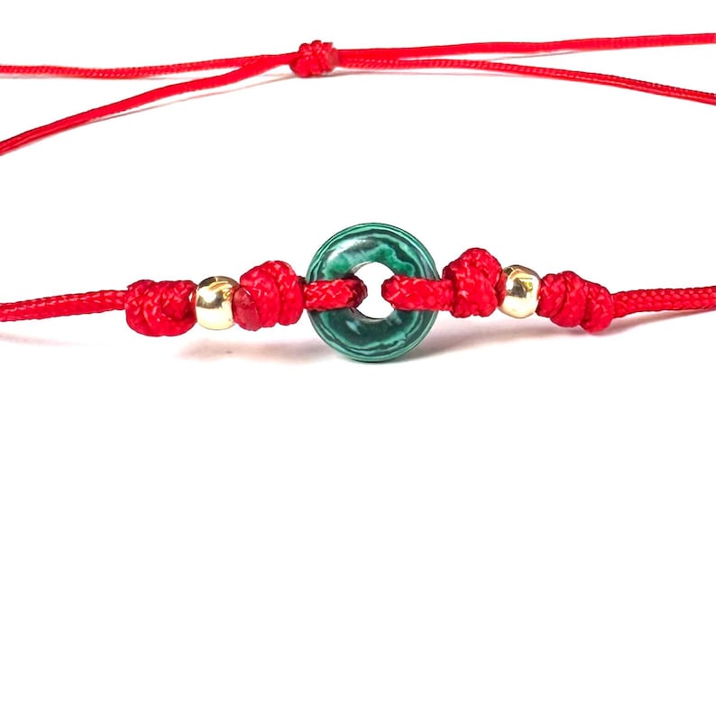 Puede incluir: Una pulsera de hilo rojo con una piedra verde circular con un patr&oacute;n &uacute;nico. La pulsera incluye cuentas doradas y nudos, sobre un fondo blanco. La cuerda est&aacute; anudada a intervalos.