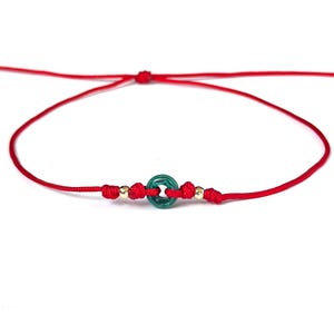 Puede incluir: Pulsera de hilo rojo con una cuenta circular verde y detalles dorados. La pulsera est&aacute; anudada y tiene un dise&ntilde;o sencillo. La pulsera se muestra sobre un fondo blanco.