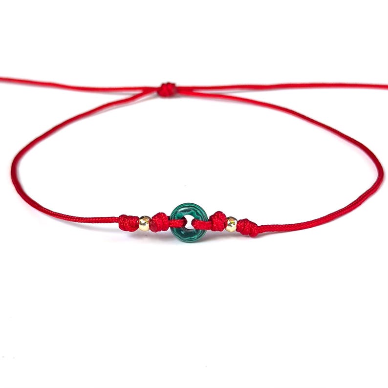 Puede incluir: Pulsera de hilo rojo con una cuenta circular verde y detalles dorados. La pulsera est&aacute; anudada y tiene un dise&ntilde;o sencillo. La pulsera se muestra sobre un fondo blanco.