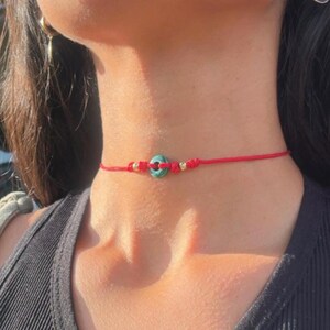 Puede incluir: Collar gargantilla rojo con una cuenta central de color turquesa y peque&ntilde;as cuentas doradas. El collar est&aacute; hecho de hilo rojo y anudado. Una pieza de joyer&iacute;a sencilla y elegante.