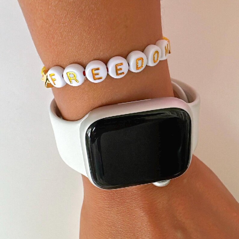 Peut inclure: Bracelet en perles blanches avec inscription dor&eacute;e "FREEDOM". Le bracelet est port&eacute; au poignet d'une personne avec une Apple Watch blanche.