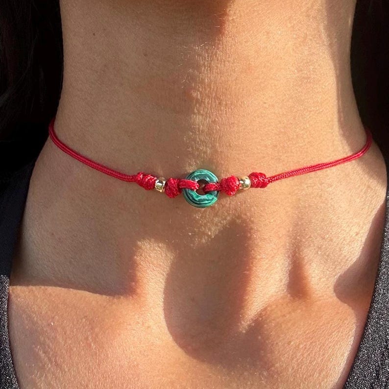Puede incluir: Un collar gargantilla de cord&oacute;n rojo con una cuenta circular verde y azul en el centro. El collar presenta cuentas doradas y nudos en el cord&oacute;n rojo. El collar se lleva alrededor del cuello.