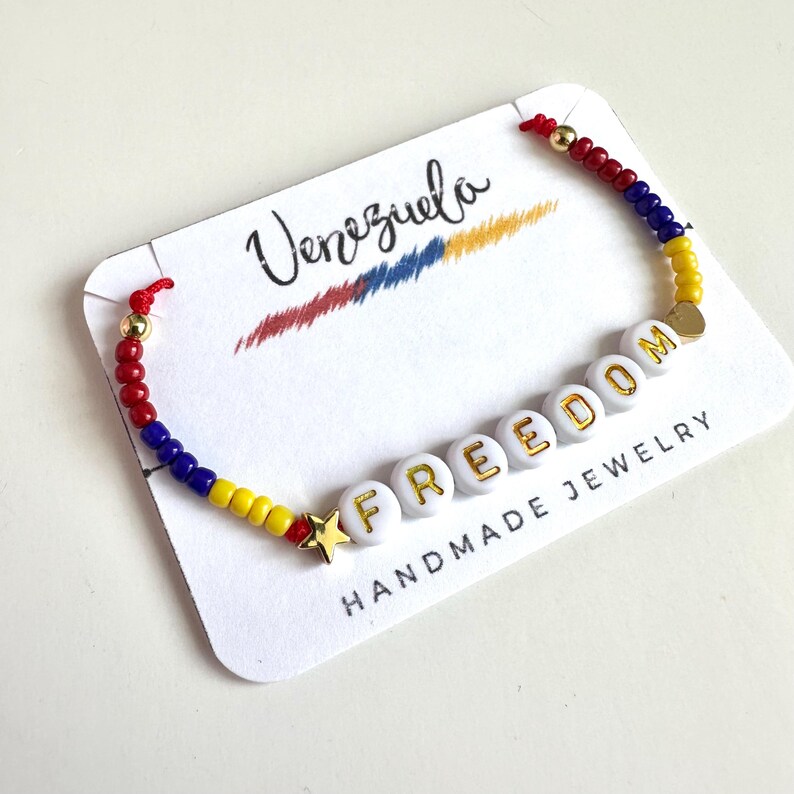 Peut inclure: Un bracelet en perles rouges, bleues et jaunes avec le mot "FREEDOM" &eacute;crit en perles blanches avec des lettres dor&eacute;es. Le bracelet est sur une carte blanche avec les mots "Venezuela" et "Bijoux faits main".