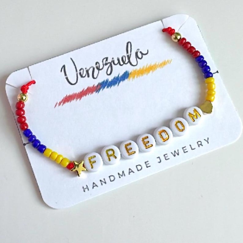 Peut inclure: Un bracelet en perles jaunes, bleues et rouges avec le mot "FREEDOM" &eacute;crit en perles blanches avec des lettres dor&eacute;es. Le bracelet est sur une carte blanche avec les mots "Venezuela" et "HANDMADE JEWELRY" &eacute;crits dessus.