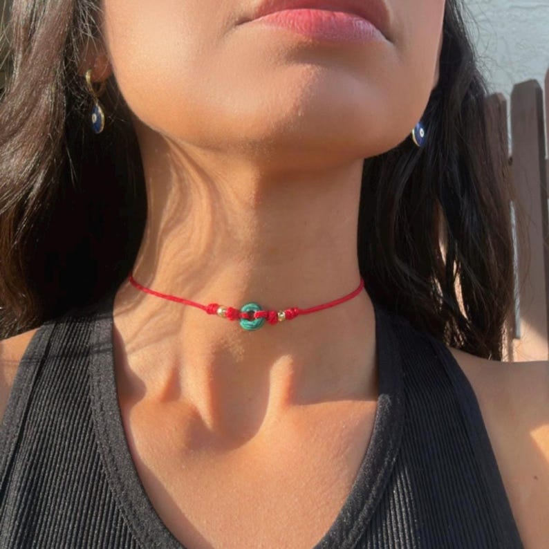 Puede incluir: Un collar gargantilla rojo con un colgante turquesa y dorado. El collar est&aacute; hecho de un fino cord&oacute;n rojo con un peque&ntilde;o colgante circular turquesa en el centro y cuentas doradas a cada lado.
