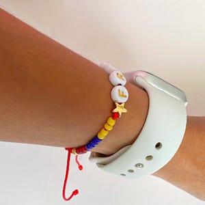 Peut inclure: Un bracelet en perles rouges, jaunes et bleues avec des perles blanches en forme de lettres qui &eacute;pellent "F E" et un charme en forme d'&eacute;toile dor&eacute;e. Le bracelet est port&eacute; au poignet d'une personne avec un bracelet de montre Apple blanc.