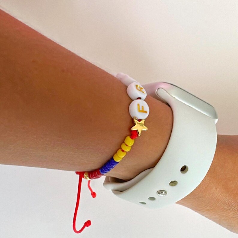 Peut inclure: Un bracelet en perles rouges, jaunes et bleues avec des perles blanches en forme de lettres qui &eacute;pellent "F E" et un charme en forme d'&eacute;toile dor&eacute;e. Le bracelet est port&eacute; au poignet d'une personne avec un bracelet de montre Apple blanc.