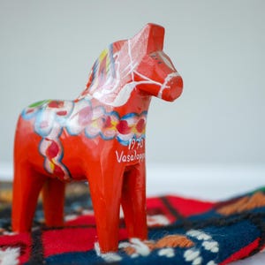 Pode incluir: Um cavalo de madeira vermelho com detalhes pintados em branco e coloridos. O cavalo é um cavalo Dala sueco tradicional, uma lembrança de arte popular. O cavalo tem o texto "1970 Vasaloppet" pintado no seu lado.