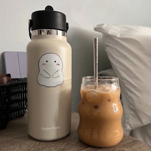 May include: A beige Hydro Flask water bottle with a ghost sticker on it. The bottle is sitting on a wooden table next to a glass of iced coffee with a metal straw.