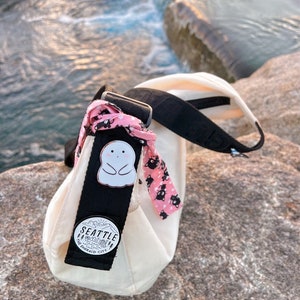 May include: A white canvas tote bag with a black strap and a black and white pin with a ghost design. The bag also has a pink and black patterned ribbon attached to the strap. The bag has a black and white pin with the text "Seattle The Emerald City" on it.