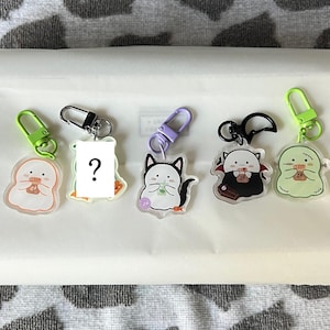 May include: A collection of five acrylic keychains with cartoon character designs. The keychains feature various characters, including a ghost, a cat, and a vampire, each with a unique design and color scheme. Each keychain has a metal clasp.