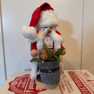 May include: A wooden nutcracker figure dressed as Santa Claus with a red velvet hat, white faux fur trim, and a white beard. The figure holds a grey fabric sack containing a small brown wooden bear and green leaves, standing on a round base.