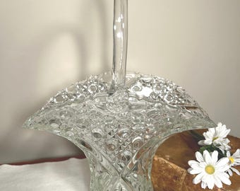 May include: Clear glass basket with a handle, featuring a textured pattern. The basket has a flared shape and a curved handle. The basket is sitting on a white surface. The basket is next to a vintage book and white flowers.