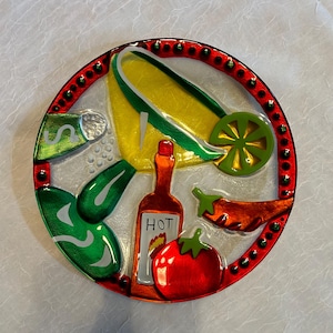 May include: A round, decorative glass plate featuring a colorful Mexican food design. The plate includes images of corn, a lime slice, a chili pepper, a tomato, a bottle of hot sauce, and a green pepper. The rim is red with small black dots.