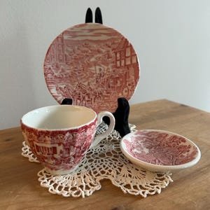 May include: A vintage tea set with a red and white transferware design. The set includes a teacup, saucer, and small plate, all featuring a detailed landscape scene. The pieces are arranged on a white lace doily.
