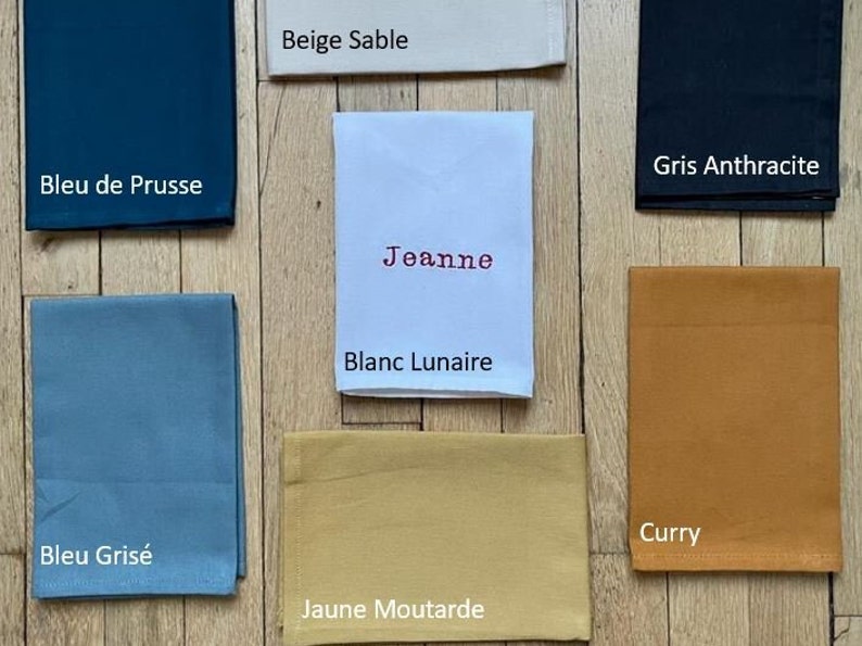 7 serviettes de table personnalisées avec écrit le nom de la couleur: bleu grisé, jaune moutarde, curry, gris anthracite, beige sable, blanc, bleu de prusse. Celle ci est brodée en blanc du prénom Ernest