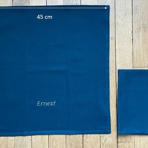une serviette bleue personnalisée, brodée du prénom Ernest. Les dimensions sont indiquées: 45cm x 45cm. A côté, la même serviette pliée