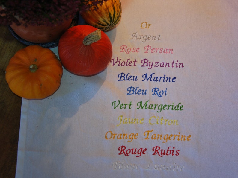 une serviette de table blanche sur laquelle 11 noms de couleurs sont brodés, correspondant aux couleurs que vous pouvez choisir pour vos broderies : or, argent, rose, violet, marine, bleu roi, vert, jaune, orange, rouge, blanc.