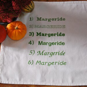 Une serviette de table blanche avec 6 fois le mot "Margeride" brodé en 6 polices différentes. Le tout est posé sur une table en bois, 2 légumes et une plante entourent le tout.