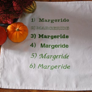 Une serviette de table blanche avec 6 fois le mot "Margeride" brodé en 6 polices différentes. Le tout est posé sur une table en bois, 2 légumes et une plante entourent le tout.