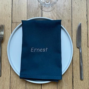 une serviette bleue, personnalisée, brodée du prénom Ernest. Elle est pliée et posée sur une assiette blanche