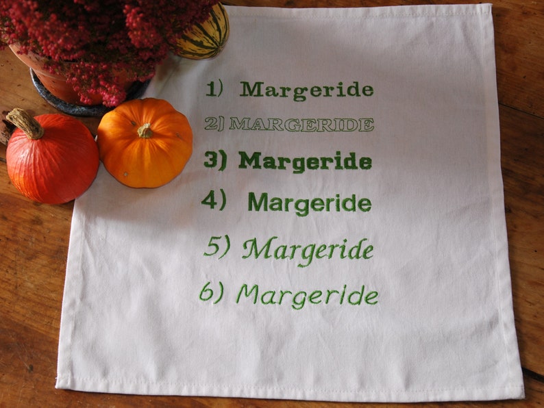 Une serviette de table blanche avec 6 fois le mot "Margeride" brodé en 6 polices différentes. Le tout est posé sur une table en bois, 2 légumes et une plante entourent le tout.