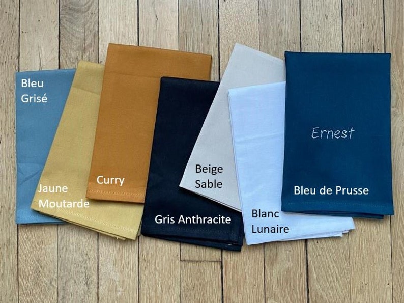 7 serviettes de table personnalisées avec écrit le nom de la couleur: bleu grisé, jaune moutarde, curry, gris anthracite, beige sable, blanc, bleu de prusse. Celle ci est brodée en blanc du prénom Ernest