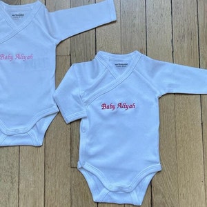 2 bodys blancs pur coton sont l un à coté de l autre, l un de taille 3 mois, l autres de tailles 6 mois. Au niveau de la poitrine du bébé, sont brodés les mots Baby Aliyah en lettres  anglaises rouges et roses.