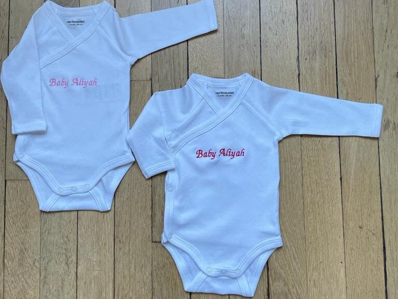 2 bodys blancs pur coton sont l un à coté de l autre, l un de taille 3 mois, l autres de tailles 6 mois. Au niveau de la poitrine du bébé, sont brodés les mots Baby Aliyah en lettres  anglaises rouges et roses.