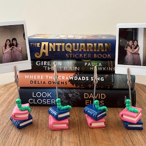 May include: Four bookends shaped like stacks of books with a green worm on top. The bookends are blue and pink. The bookends are holding up instant photos of two women.