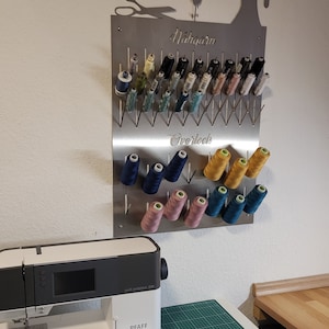 Könnte beinhalten: Ein Wandorganizer aus Metall mit einer Nähmaschinen-Silhouette, beschriftet mit "Nähgarn" und "Overlock". Er hat Reihen von Haken zum Aufbewahren von Garnrollen in verschiedenen Farben, darunter Rosa, Blau, Gelb und Grün.