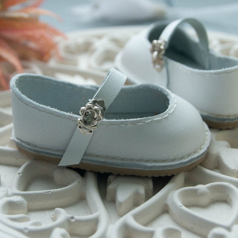 May include: A pair of white leather baby shoes with silver buckles. The shoes have a simple design and are perfect for a special occasion.