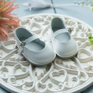 May include: A pair of white leather doll shoes with silver buckles. The shoes are on a white decorative plate with a heart design.