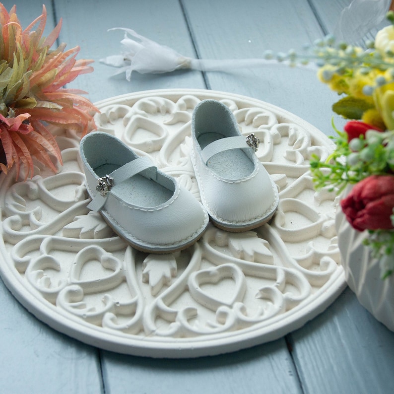May include: Two white leather baby shoes with silver buckles on a white decorative plate with a heart design.