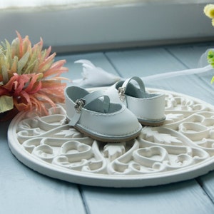 May include: A pair of white leather baby shoes with silver buckles, sitting on a white decorative plate with a floral design.