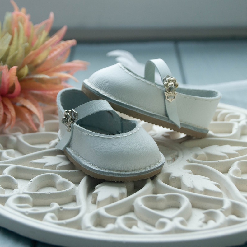 May include: A pair of white leather baby shoes with a strap and buckle detail. The shoes are on a white decorative surface.