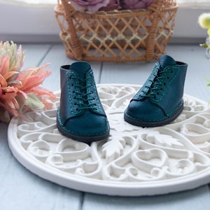 May include: A pair of teal-colored miniature boots with green laces and stitching. The boots are made of textured material and sit on a white, ornate, circular decorative piece. The boots are a small size, suitable for dolls or other small figures.