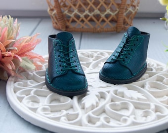 May include: Two teal miniature boots with green laces and stitching. The boots are made of textured material and are displayed on a white, ornate, circular decorative piece. The boots are a small size, suitable for dolls or other small figures.