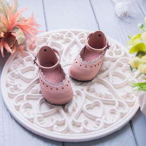 May include: A pair of pink leather doll shoes with white lace trim and a buckle closure. The shoes are sitting on a white decorative plate with a heart design.