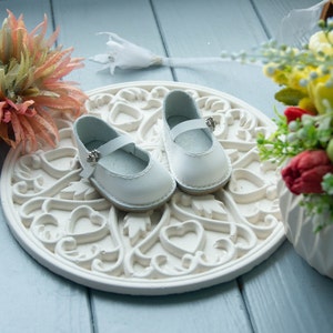 May include: A pair of white leather baby shoes with a buckle closure, displayed on a white decorative plate with a heart design.