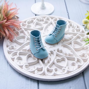 Może przedstawiać: Para małych, jasnoniebieskich skórzanych butów ze sznurowadłami. Buty są prezentowane na białym, ozdobnym, okrągłym talerzu. Buty mają około 5 cm wysokości. Talerz ma szczegółowy kwiatowy wzór.