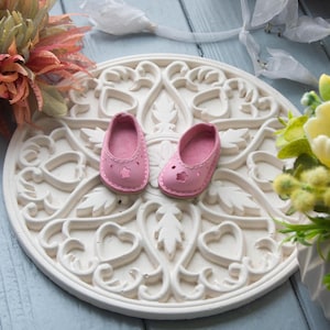 May include: A pair of pink leather baby shoes with a flower cutout design on a white decorative plate.