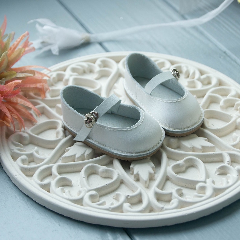 May include: A pair of white leather doll shoes with silver buckles. The shoes are sitting on a white decorative plate with a heart design.