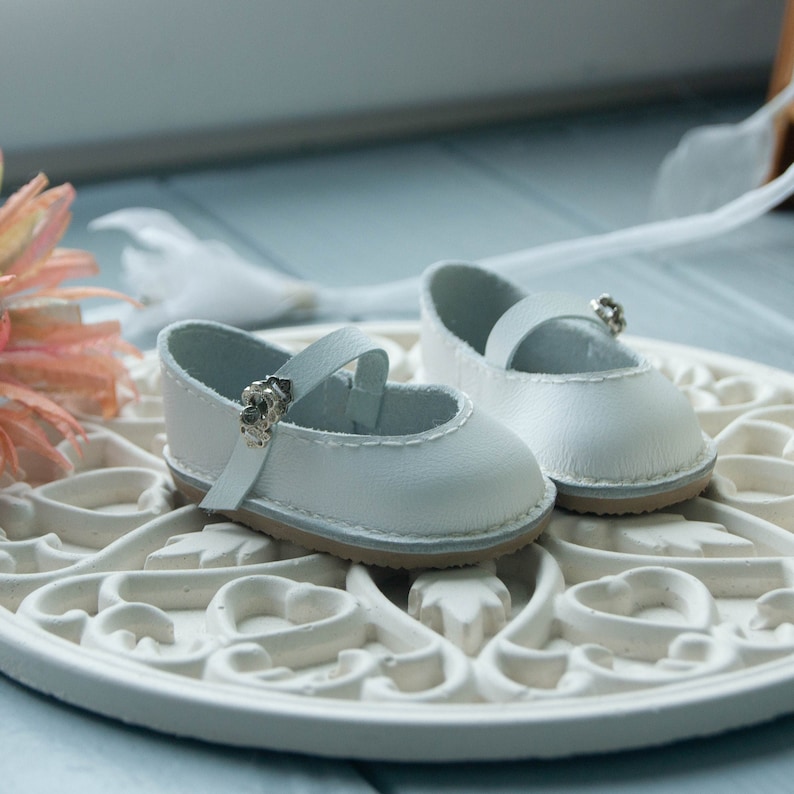 May include: A pair of white leather baby shoes with silver buckles. The shoes are on a white decorative plate with a floral design.