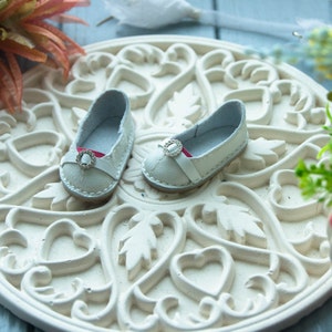 May include: Two white leather doll shoes with a pink ribbon and a silver buckle on a white ornate background.